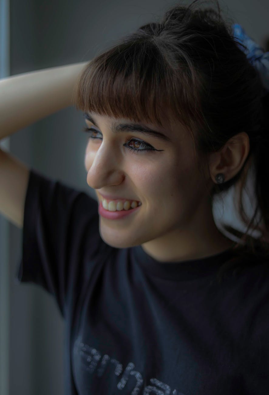 Indie Sleaze Fringes: The Messy, Cool Girl Bangs 1 young woman smiling by a window in soft light